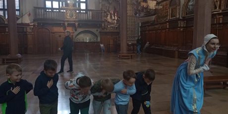 Powiększ grafikę: Panienka z Okienka pokazuje dzieciom w jaki sposób należy składać pokłon. Dzieci dostojnie pochylają się.