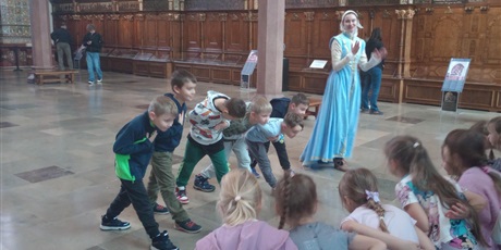 Powiększ grafikę: Panienka z Okienka pokazuje dzieciom w jaki sposób należy składać pokłon. Dzieci dostojnie pochylają się.