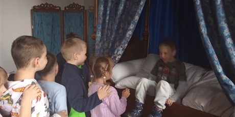 Powiększ grafikę: Chłopiec próbuje usiąść na łóżku z baldachimem. Śmieje się przy tym. Łóżko jest drewniane i pokryte dużą ilością pościeli.