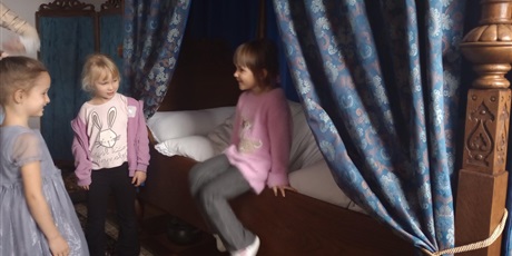 Powiększ grafikę: Dziewczynka próbuje usiąść na łóżku z baldachimem. Śmieje się przy tym. Łóżko jest drewniane i pokryte dużą ilością pościeli.