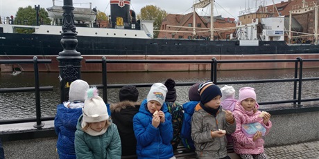 Powiększ grafikę: Dzieci ubrane w ciepłe kurtki zajadają bułeczki. W tle widać statek Sołdek.