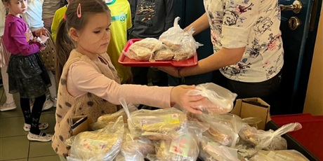 Powiększ grafikę: Grupa dzieci przekłada samodzielnie przygotowane kanapki z tacy do pojemnika. Pojemnik zostanie zawieziony do potrzebujących.