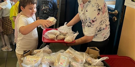 Powiększ grafikę: Grupa dzieci przekłada samodzielnie przygotowane kanapki z tacy do pojemnika. Pojemnik zostanie zawieziony do potrzebujących.