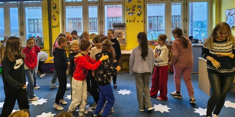 Powiększ grafikę: Grupa dzieci wraz z  nauczycielką tańczy . Na podłodze między nimi leżą białe papierowe gwiazdy.