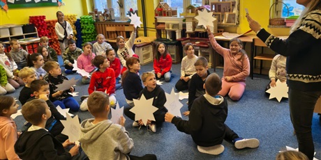 Powiększ grafikę: Grupa dzieci siedzi na dywanie w świetlicy. Część z nich trzyma w ręku białe papierowe gwiazdy. Dwoje dzieci unoszą je do góry. Nauczycielka z prawej strony czyta wróżbę z kartki.