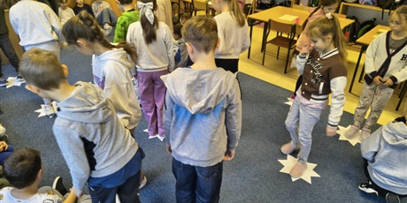 Powiększ grafikę: Grupa dzieci  tańczy . Na podłodze między nimi leżą białe papierowe gwiazdy.