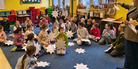 Powiększ grafikę: Grupa dzieci siedzi na dywanie w świetlicy. Część z nich trzyma w ręku białe papierowe gwiazdy. Dziewczynka  unosi ją do góry. Nauczycielka z prawej strony czyta wróżbę z kartki.