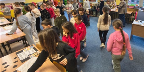 Powiększ grafikę: Dzieci w świetlicy w trakcie zabawy andrzejkowej. Część dzieci chodzi po dywanie lub stoi w kolejce. Część z nich bawi się korzystając z wróżb, które znajdują się na stołach.