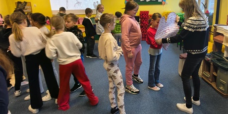 Powiększ grafikę: Dzieci w świetlicy w trakcie zabawy andrzejkowej. Część dzieci chodzi po dywanie lub stoi w kolejce do nauczycielki, która trzyma w ręku duże papierowe serce.