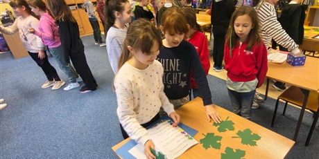 Powiększ grafikę: Dzieci w świetlicy w trakcie zabawy andrzejkowej. Część dzieci chodzi po dywanie lub stoi w kolejce. Trzy dziewczynki stoją przy stole , na którym znajdują się zielone liście z wróżbami.