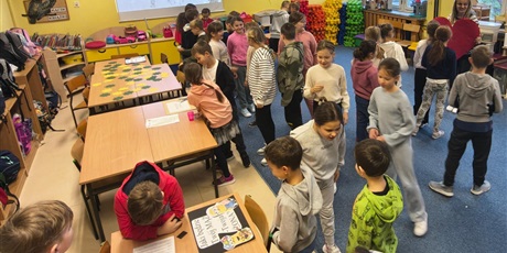 Powiększ grafikę: Dzieci w świetlicy w trakcie zabawy andrzejkowej. Część dzieci chodzi po dywanie lub stoi w kolejce. Część z nich bawi się korzystając z wróżb, które znajdują się na stołach.