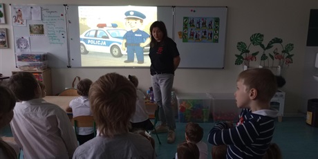 Powiększ grafikę: Pani pokazuje dzieciom filmik, na którym widać policjanta. Dzieci w skupieniu słuchają.