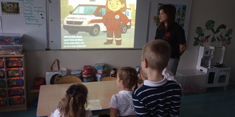 Powiększ grafikę: Pani pokazuje dzieciom filmik, na którym widać karetkę pogotowia. Dzieci w skupieniu słuchają.