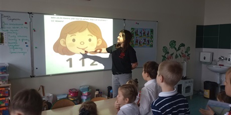 Powiększ grafikę: Pani pokazuje dzieciom filmik, na którym widać numer alarmowy 112. Dzieci w skupieniu słuchają. Starają się zapamiętać ten numer.
