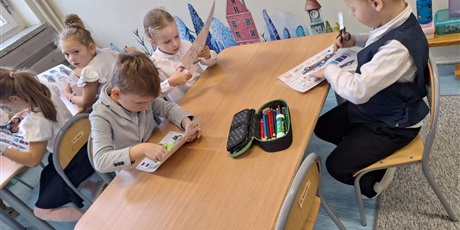 Powiększ grafikę: Dzieci siedzą przy stolikach i wykonują pracę plastyczną. Wycinają nożyczkami z kartek z papieru.