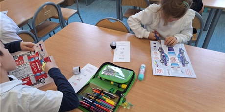 Powiększ grafikę: Dzieci siedzą przy stolikach i wykonują pracę plastyczną. Wycinają nożyczkami z kartek z papieru.