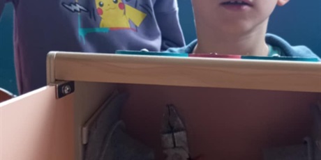 Powiększ grafikę: Na zdjęciu widać chłopca w niebieskiej bluzie z rysunkiem Pikachu, stojącego przy oknie i spoglądającego na kolegę siedzącego przed nim. Siedzący chłopiec trzyma ręce w otworach drewnianego pudełka,