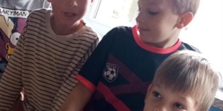 Powiększ grafikę: Zabawa w rozpoznawanie przedmiotów trwa. Trzech chłopców stojących na tle jasnego okna przygląda się koledze, który ma do rozpoznania  szpachelkę umieszczoną w drewnianym pojemniku.