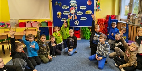 Powiększ grafikę: Grupa kilkunastu chłopców siedzi na dywanie w świetlicy. Część z nich ma uniesione ręce do góry. Z tyłu znajduje się granatowa tablica z dekorację z okazji Dnia Chłopaka.