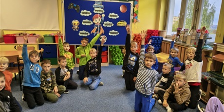 Powiększ grafikę: Grupa kilkunastu chłopców siedzi na dywanie w świetlicy. Część z nich ma uniesione ręce do góry. Z tyłu znajduje się granatowa tablica z dekorację z okazji Dnia Chłopaka.