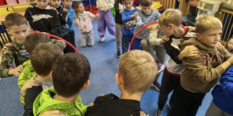 Powiększ grafikę: Kilkunastu chłopców stoi w dwóch rzędach trzymając się za ramiona. Każdy rząd przekłada hula hop nie odrywając rąk. Z tyłu znajdują się dwie dziewczynki.