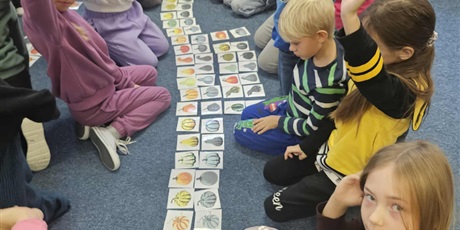 Powiększ grafikę: Grupa dzieci siedzi na dywanie w świetlicy. Miedzy nimi na podłodze leżą karty z kolorowymi dyniami i ich cieniami. Kilkoro dzieci ma poniesiona rękę do góry.