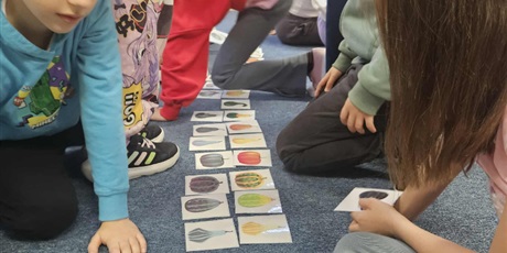 Powiększ grafikę: Grupa dzieci siedzi na dywanie w świetlicy. Miedzy nimi na podłodze leżą karty z kolorowymi dyniami i ich cieniami.  Dzieci układają te karty.
