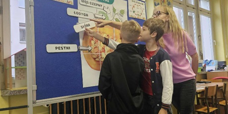 Powiększ grafikę: Dwóch chłopców zawiesza na granatowej tablicy  nazwy elenentów budowy dyni. Pomaga im nauczycielka.