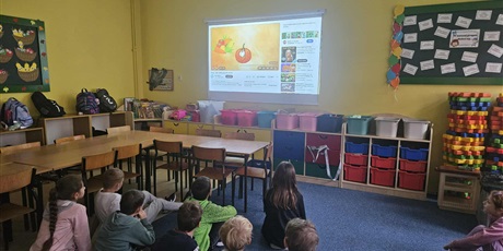 Powiększ grafikę: Grupa dzieci siedzi na dywanie i wpatruje się w ekran ścienny, na którym jest wyświetlany film o pochodzeniu dyni.