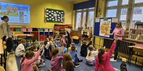 Powiększ grafikę: Grupa dzieci siedzi na dywanie w świetlicy zwrócona w stronę tablicy, przy której stoi nauczycielka mówiąca do dzieci. Kilkoro dzieci ma poniesiona rękę do góry.