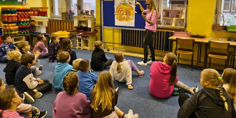 Powiększ grafikę: Grupa dzieci siedzi na dywanie w świetlicy zwrócona w stronę tablicy, przy której stoi nauczycielka mówiąca do dzieci.