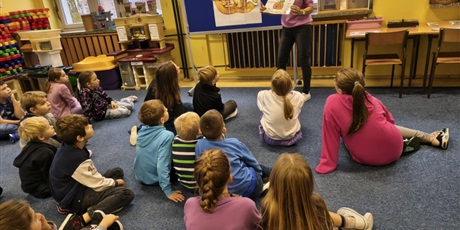 Powiększ grafikę: Grupa dzieci siedzi na dywanie w świetlicy zwrócona w stronę tablicy, przy której stoi nauczycielka trzymająca w ręku planszę.