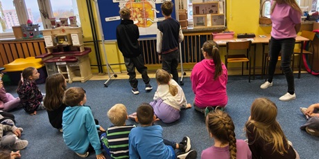 Powiększ grafikę: Dwóch chłopców zawiesza na granatowej tablicy nazwy elementów budowy dyni. Obok stoi nauczycielka. Na dywanie siedzą dzieci wpatrujące się w tablicę.