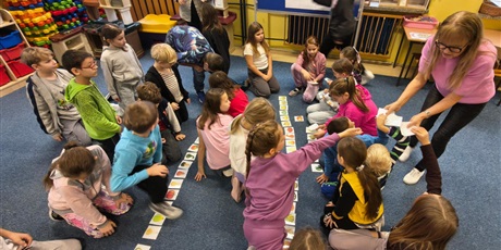 Powiększ grafikę: Duża grupa dzieci układa na dywanie karty przedstawiające różne rodzaje dyni i jej cieni. Dwie nauczycielki są wśród dzieci i podają im karty.