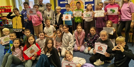 Powiększ grafikę: Grupa dzieci wraz z dwoma nauczycielkami pozuje do zdjęcia. W rękach trzymają karty z elementami dyni. W tle stoi granatowa tablica informacyjna zawierająca opis budowy dyni.