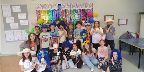 Powiększ grafikę: Grupa dzieci z klasy 2C pozuje do zdjęcia. Na głowach mają peruki, kapelusze oraz kolorowe okulary.  Kilkoro dzieci z przodu siedzi na podłodze, pozostali stoją z tyłu.