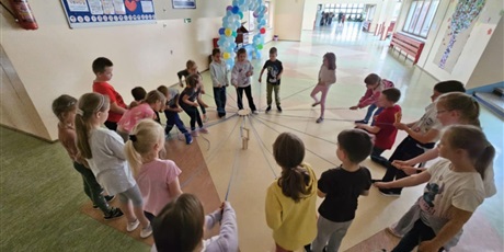Powiększ grafikę: Grupa dzieci z klasy 1B stoi na korytarzu szkolnym. Dzieci ustawione są w kole. Każdy w ręku trzyma niebieski sznurek.