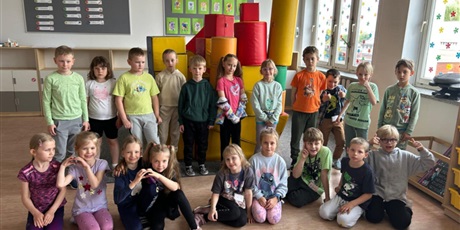 Powiększ grafikę:  Grupa dzieci z klasy 1C pozuje do zdjęcia. Część z nich siedzi na podłodze, reszta stoi z tyłu. Za dziećmi widoczne są tablice z napisami i rysunkami. Stoi również wieża zbudowana z kolorowych klockó
