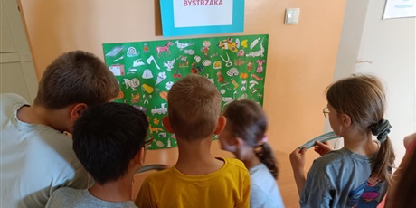 Powiększ grafikę: Grupa dzieci stoi tyłem do aparatu. Wpatrują się w zieloną planszę z rysunkami, która wisi przed nimi na ścianie.