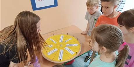 Powiększ grafikę: Dzieci i wolontariuszka  pochylają się nad  kołem fortuny, które leży na stole. Na ścianie wisi kartka z napisem TEST SPORTOWCA.