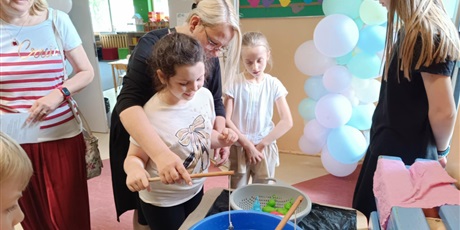 Powiększ grafikę:  Dziewczynka z pomocą nauczycielki łowi kolorowe kaczki. W ręku dziewczynka trzyma drewnianą wędkę. Obok stoją dwie uczennice i nauczycielka.