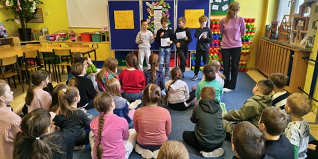 Powiększ grafikę: Grupa dzieci siedzi na dywanie w świetlicy tyłem do obiektywu. Czterech chłopców stoi na tle granatowej tablicy, w ręku trzymają białe kartki. Obok stoi nauczycielka.