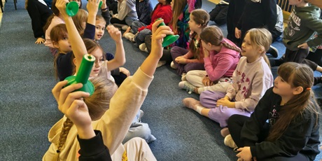 Powiększ grafikę: Dzieci siedzą na dywanie w dwóch rzędach na przeciwko siebie. Część z nich ma uniesione ręce do góry. Między nimi na podłodze stoją zielone słupki.