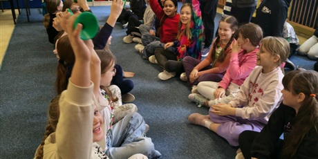 Powiększ grafikę: Dzieci siedzą na dywanie w dwóch rzędach na przeciwko siebie. Część z nich ma uniesione ręce do góry. Między nimi na podłodze stoją zielone słupki.