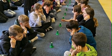 Powiększ grafikę: Dzieci siedzą na dywanie w dwóch rzędach na przeciwko siebie. Część z nich ma uniesione ręce do góry. Między nimi na podłodze stoją zielone słupki.