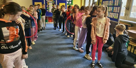 Powiększ grafikę: Dzieci stoją w dwóch rzędach trzymając się za ramiona. Oba rzędy próbują przejść przez hula hop bez odrywania rąk.