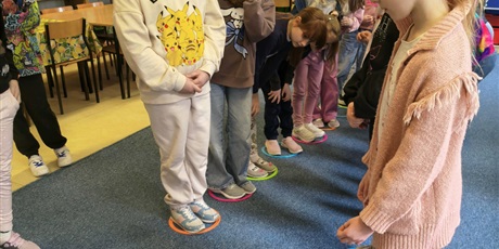 Powiększ grafikę: Dzieci stoją w dwóch rzędach na przeciwko siebie. Każdy stoi w kolorowym kółku. Na podłodze leży zielona kostka do gry.