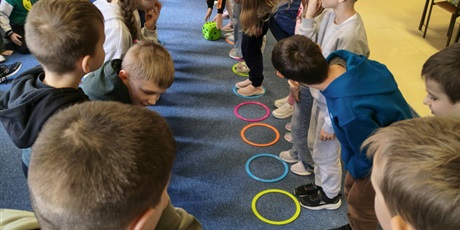 Powiększ grafikę: Dzieci stoją w dwóch rzędach na przeciwko siebie. Część z nich stoi w kolorowym kółku. Na podłodze leży zielona kostka do gry.