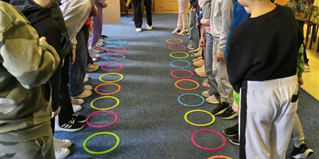 Powiększ grafikę: Dzieci stoją w dwóch rzędach na przeciwko siebie. Na podłodze leżą kolorowe kółka. Z tyłu stoi nauczycielka, która trzyma w ręku zieloną kostkę do gry i tłumaczy zasady rozgrywki.