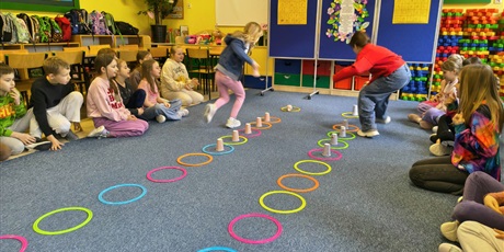 Powiększ grafikę: Dzieci siedzą na dywanie w dwóch rzędach na przeciwko siebie. Między nimi ułożone są dwa rzędy kolorowych kółek z kubkami w środku. Dwie dziewczynki rywalizują składając pojedynczo kubki.